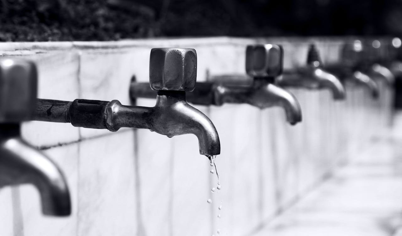 expertise-01 Black and white image of a row of faucets with water dripping, highlighting plumbing details.
